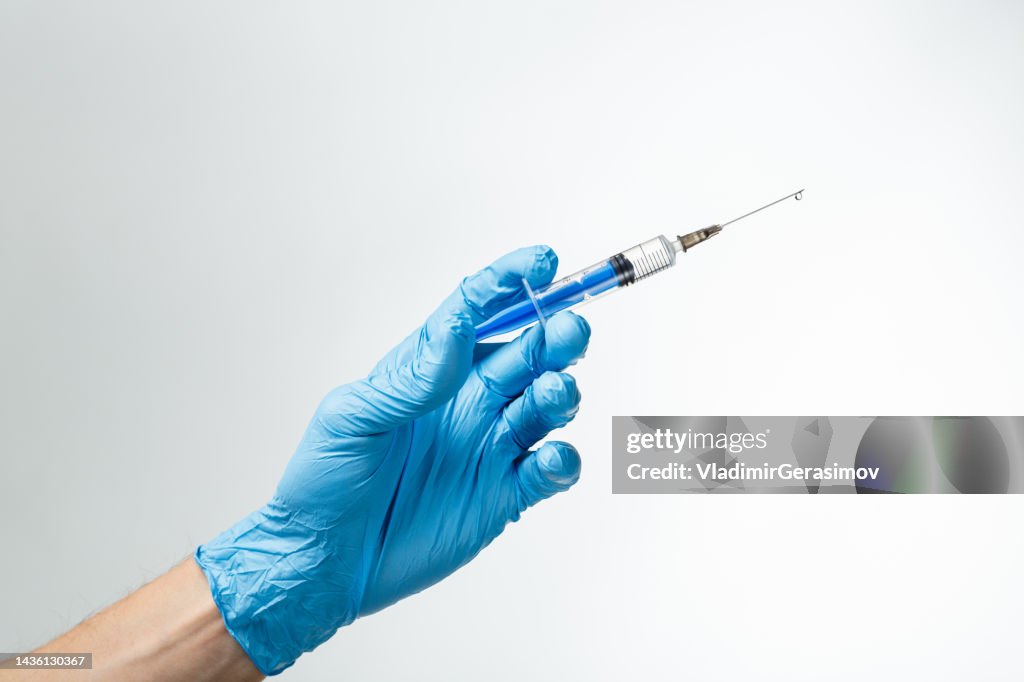 Syringe with a drop on a needle in hand in a blue rubber glove on a white background.