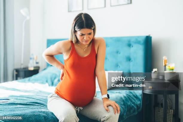 pregnant young woman sitting on the bed experiencing pain in the lower back - labour childbirth stock pictures, royalty-free photos & images