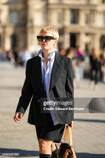 Guest wears black sunglasses, gold earrings, a pale blue ruffled high neck blouse, a black blazer jacket, black suit shorts, black shiny leather...