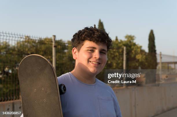 portrait of smiling teenage boy holding skateboard looking at camera. close up of teenage skateboarder holding his skateboard. - skater boy hair stock pictures, royalty-free photos & images