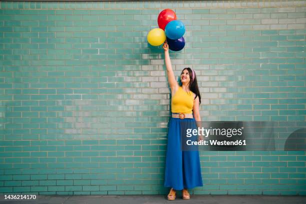 woman with hand raised holding multi colored balloons in front of teal brick wall - person standing infront of wall stockfoto's en -beelden