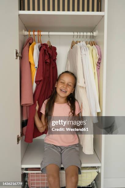 happy girl with eyes closed sitting by clothes in closet - childrens closet stock pictures, royalty-free photos & images