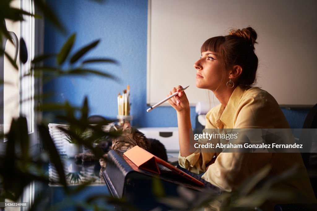 A graphic designer working from home with her drawing tablet holding her digitized pen looking away while thinking