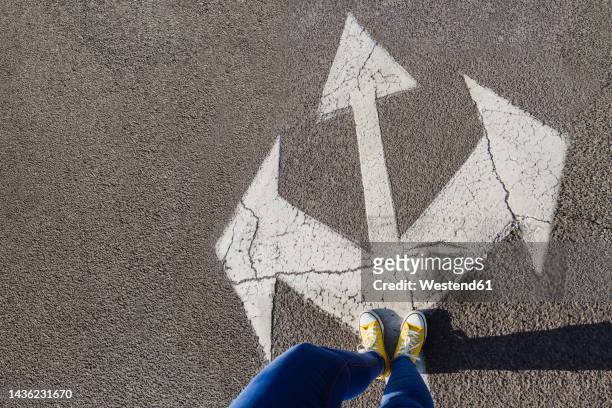 woman standing on three way direction arrow sign on road - low section of woman standing in front of arrow sign on road stock pictures, royalty-free photos & images