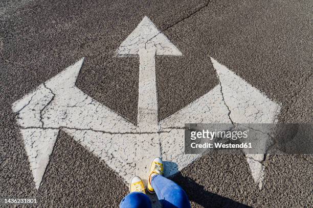 woman standing on asphalt with three way direction arrow sign - low section of woman standing in front of arrow sign on road stock pictures, royalty-free photos & images