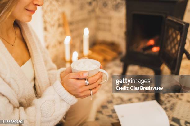 woman's hands holding cup of cocoa with marshmallows. fireplace with candles on background - haardvuur stockfoto's en -beelden