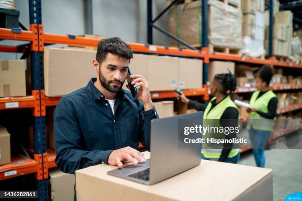 supervisore del magazzino durante una telefonata e usando il suo laptop mentre le operaie scansionano le scatole in background - industrial laborer foto e immagini stock