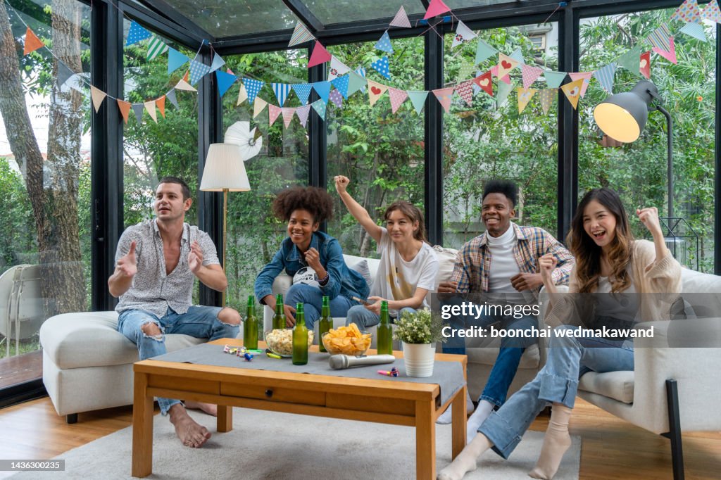 Cheerful friends sitting on the couch and exulting while watching a sport match on tv - Happy young cool people having fun at a home party
