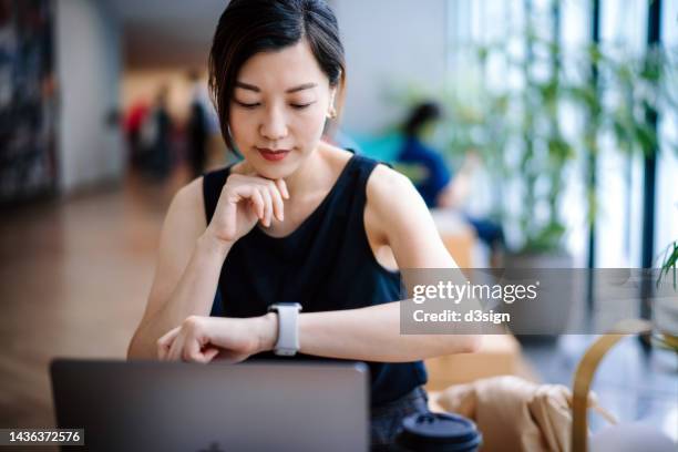 young asian businesswoman sitting at her desk in a modern office space, looks down at her watch checking the time while working on laptop. business and time management concept - instrument de mesure du temps photos et images de collection