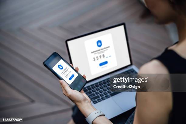 over the shoulder view of young woman using mobile device with two-factor authentication (2fa) security while logging in securely to her laptop. privacy protection, internet and mobile security - computer security stock pictures, royalty-free photos & images