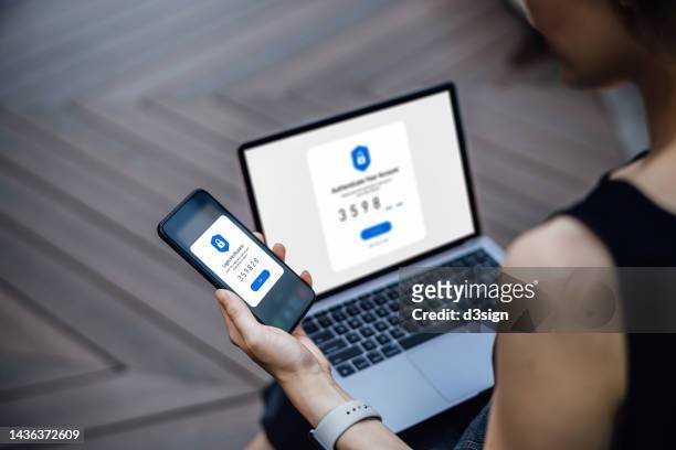 over the shoulder view of young woman using mobile device with two-factor authentication (2fa) security while logging in securely to her laptop. privacy protection, internet and mobile security - information security imagens e fotografias de stock