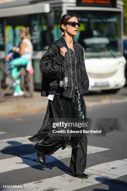 Guest wears black sport sunglasses, silver earrings, a black nylon / zipper oversized bomber coat, a black shiny leather Cagole shoulder bag from...