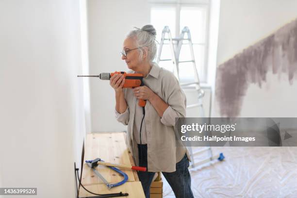 senior woman drilling into wall, preparation for hanging pictures. - rebuilding stock pictures, royalty-free photos & images