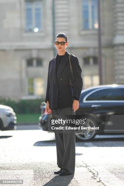 Guest wears black sunglasses, a black t-shirt, a black blazer jacket, a black shiny leather shoulder bag from Chanel, dark gray large pants, black...