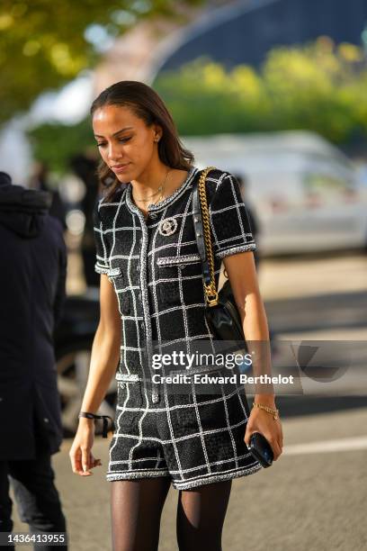 Guest wears gold earrings, a gold chain necklaces, a black and white checkered print pattern tweed short jumpsuits from Chanel, a black shiny leather...