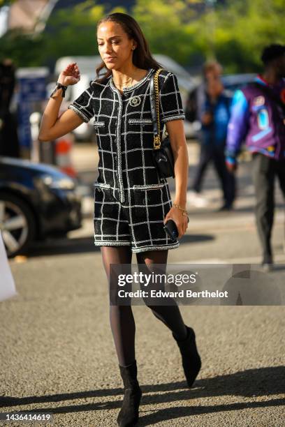 Guest wears gold earrings, a gold chain necklaces, a black and white checkered print pattern tweed short jumpsuits from Chanel, a black shiny leather...