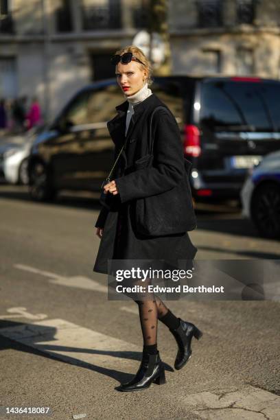 Guest wears black sunglasses, silver and diamonds earrings, a white latte ribbed wool turtleneck pullover, a black long coat, a black ribbed velvet...