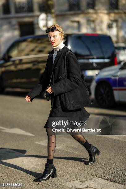 Guest wears black sunglasses, silver and diamonds earrings, a white latte ribbed wool turtleneck pullover, a black long coat, a black ribbed velvet...