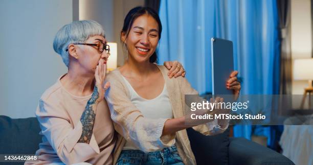 happy asian family, young daughter and senior elderly mom teach older mother to use digital tablet video call online to friends spend time in home weekend at night. - mother daughter webcam stockfoto's en -beelden