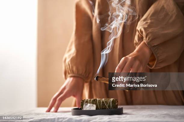the hands of a girl in a beige dress hold a smoking palo santo stick against the background of ritual things: a rock crystal stone and a twist for fumigation. - wierook gefabriceerd object stockfoto's en -beelden