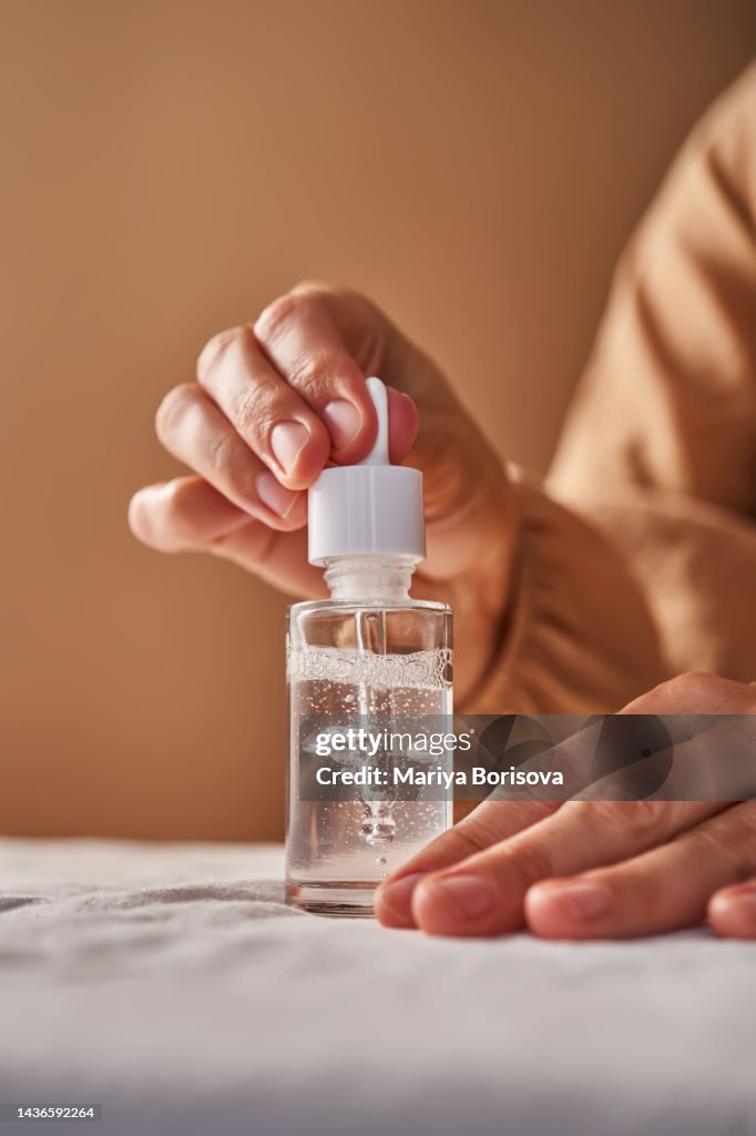 A girl in a beige dress opens a transparent bottle of serum.