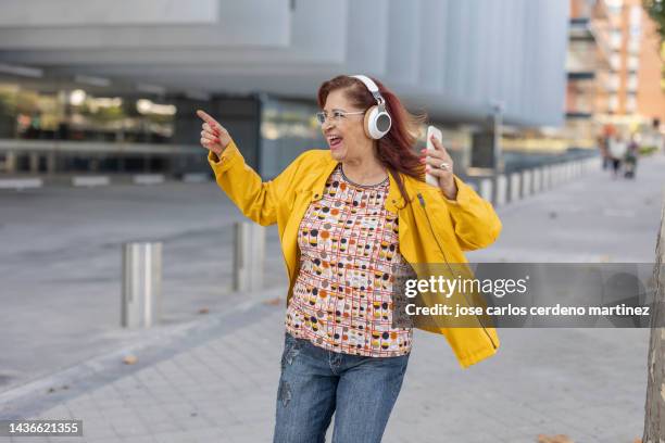 elderly curvy hispanic woman dancing with music playlist app on smartphone while wearing wireless headphones around the city. - fat woman dancing stockfoto's en -beelden