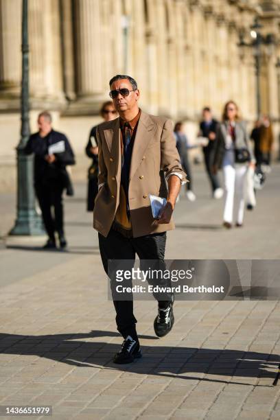 Guest wears brown sunglasses, a brown shirt, a black tie, a brown buttoned blazer jacket, black suit pants, black and white shiny leather sneakers ,...