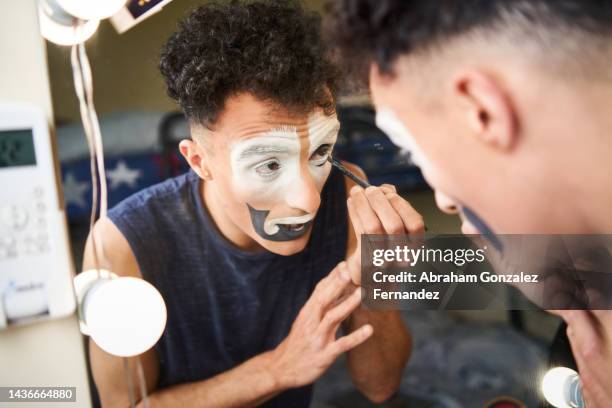 a young circus performer painting his face with a black brush while looking in the mirror in his dressing room - circus dressing room stock pictures, royalty-free photos & images