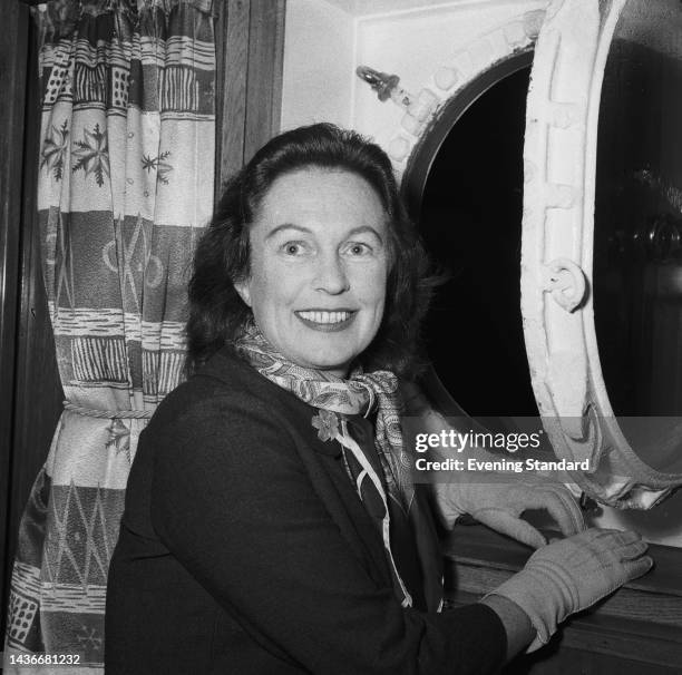 Irish actress Geraldine Fitzgerald wearing a dark jacket with gloves and a scarf around her neck, poses alongside an open porthole on an ship, 12th...