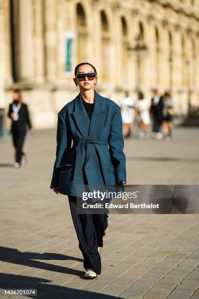 Guest wears black sunglasses, gold earrings, a black wool pullover, a navy blue oversized / belted blazer coat, black large flowing pants, beige...