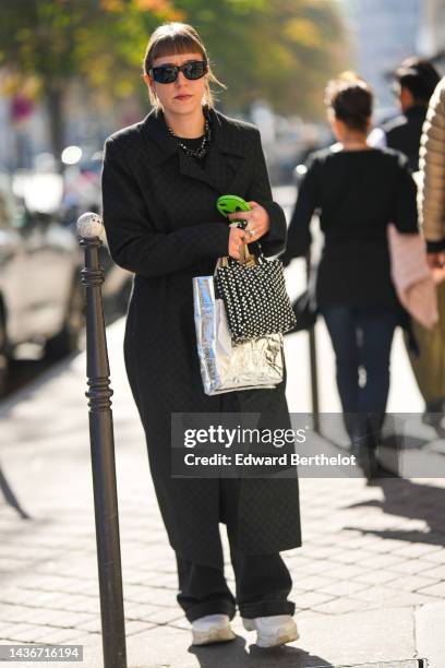 Guest wears black sunglasses, a black t-shirt, a black print pattern long coat, silver rings, black large pants, white leather and fabric sneakers, a...