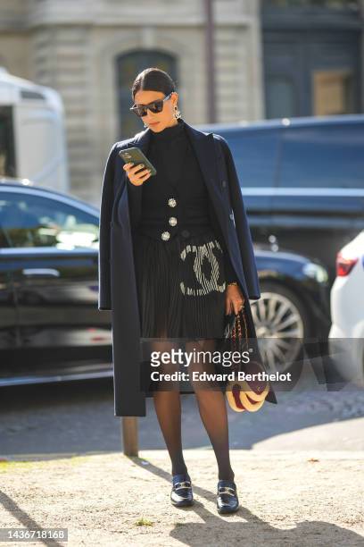 Guest wears black sunglasses, a black turtleneck t-shirt, a black large crystal buttons wool cardigan from Chanel, a navy blue long coat, a black...
