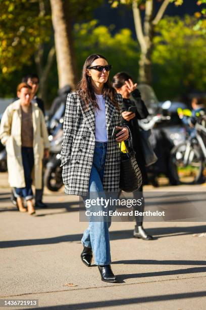 Guest wears black sunglasses, a white t-shirt, a black and white checkered print pattern tweed blazer jacket, blue faded denim large pants, black...