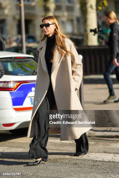 Guest wears black square sunglasses, a black wool turtleneck pullover, a beige long wool coat, black large flowing pants, black shiny leather pointed...