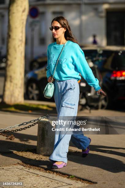 Guest wears brown sunglasses, a flashy blue with embroidered logo wool pullover from Chanel, a blue green shiny leather crossbody bag, blue denim...