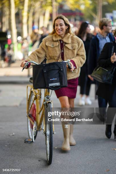 Guest wears gold earrings, a beige fur oversized bomber coat, a burgundy tweed short dress , a beige matte rain boots from Chanel , outside Chanel,...