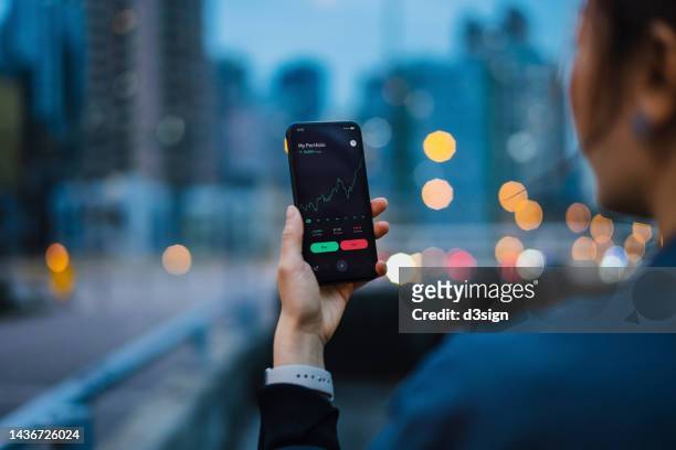 over the shoulder view of asian woman managing finance and investment, analyzing stock market data with mobile app on smartphone in the city at dusk. stock exchange, banking, finance, investment, financial trading concept. smart banking with technology - fintech imagens e fotografias de stock