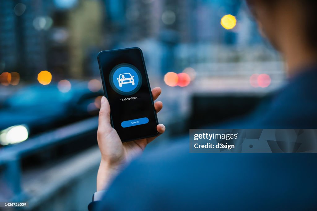 Over the shoulder view of young Asian woman arranging a taxi service with mobile app on smartphone in city street in the evening, with illuminated city scene in background. Lifestyle and transportation technology. Convenience and trustworthy car service