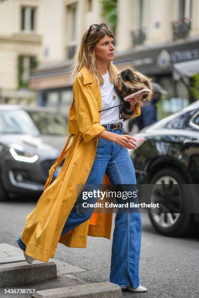 Guest wears brown sunglasses, gold earrings, a white t-shirt, a yellow long trench coat, a black shiny leather with gold buckle belt from Celine,...
