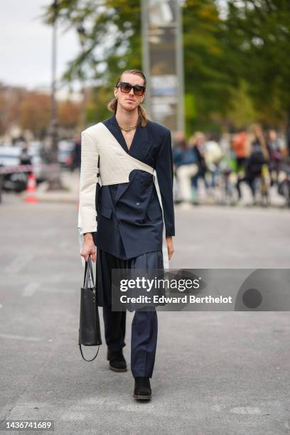 Guest wears black sunglasses, gold earrings, a navy blue blazer jacket, a navy blue pleated / accordion knees skirt, matching navy blue suit pants, a...