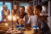 Happy Jewish boy and his grandfather lighting the menorah during family meal on Hanukkah.