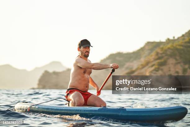 man with a cap sitting on the paddle board while paddleboarding in the sea. - paddle board men imagens e fotografias de stock