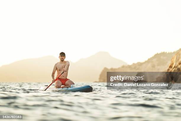 man in cap sitting on paddle board while rowing in the sea. - paddle board men imagens e fotografias de stock