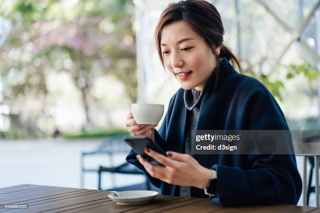 Smiling young Asian woman sitting in an outdoor cafe, having a cup of coffee and text messaging on smartphone. Enjoying a relaxing afternoon and carefree moment. Lifestyle and technology