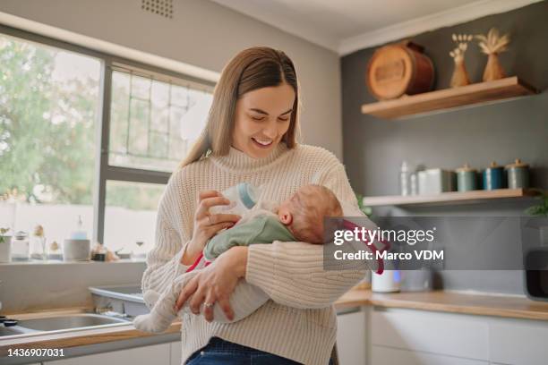 madre che nutre il suo bambino con una bottiglia di latte con amore, cura e felicità mentre sta in cucina. felice, sorridente e neonato bambino che beve latte artificiale da sua madre nella loro moderna casa di famiglia. - woman smile kitchen foto e immagini stock