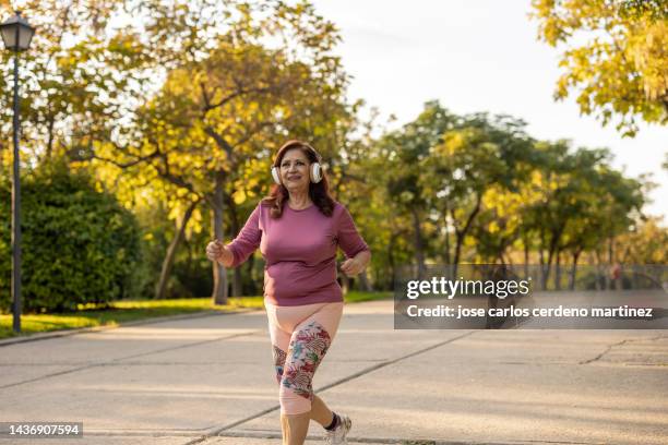 aged sportswoman jogging in the park while listening to music at sunset - big fat white women stock-fotos und bilder