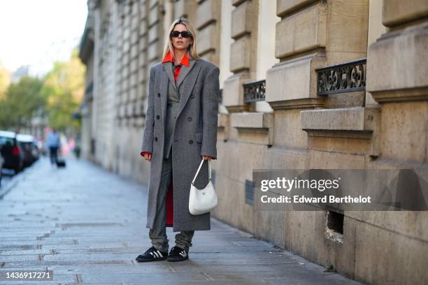 Natalia Verza wears black sunglasses, gold earrings, a neon orange shirt, a pale gray buttoned long coat, a gray oversized long coat, gray wool large...