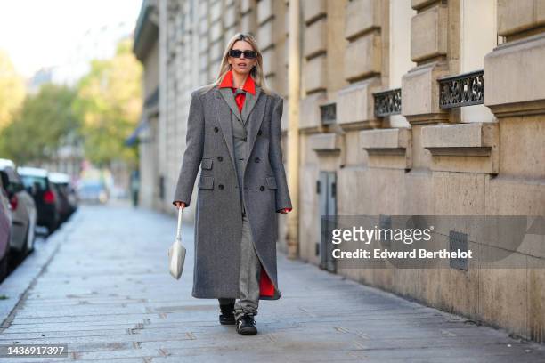 Natalia Verza wears black sunglasses, gold earrings, a neon orange shirt, a pale gray buttoned long coat, a gray oversized long coat, gray wool large...