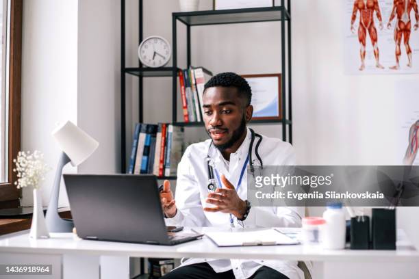 african young doctor is wearing a lab coat and a stethoscope discussing on video call over laptop while sitting at clinic. - online doctor stock pictures, royalty-free photos & images