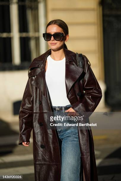 Diane Batoukina wears black sunglasses, a white ribbed cropped tank-top, a dark brown shiny varnished leather long coat, a black grained leather...